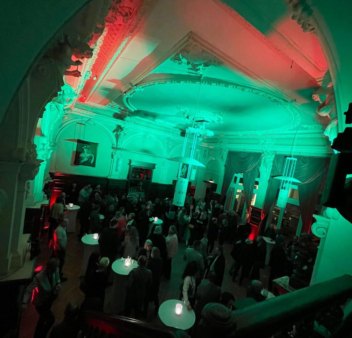 Gäste an Stehtischen in der Josephine Bar, der Raum ist in Türkis und Rot ausgeleuchtet, darüber die historische Stuckdecke